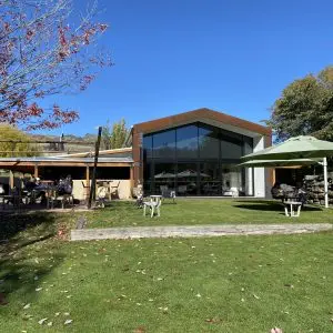 Modern building with large windows on grassy lawn, tables with umbrellas, trees, mountains visible.